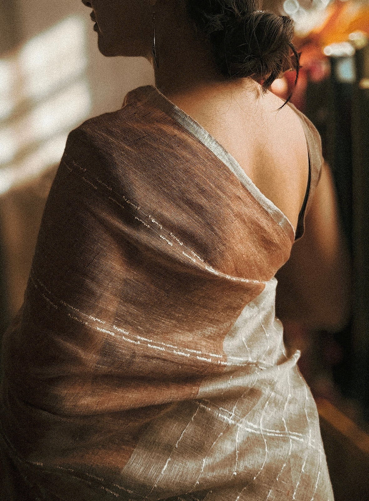 An image of woman wearing a dusky brown linen saree and is standing sideways with her pallu stretched towards the front. 