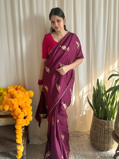 Woman in a purple saree with gold patterns standing indoors with decorative elements.