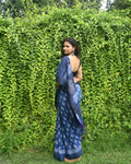 Woman wearing a blue saree with a pattern, standing against a green hedge.