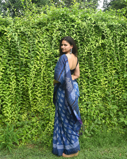 Woman wearing a blue saree with a pattern, standing against a green hedge.