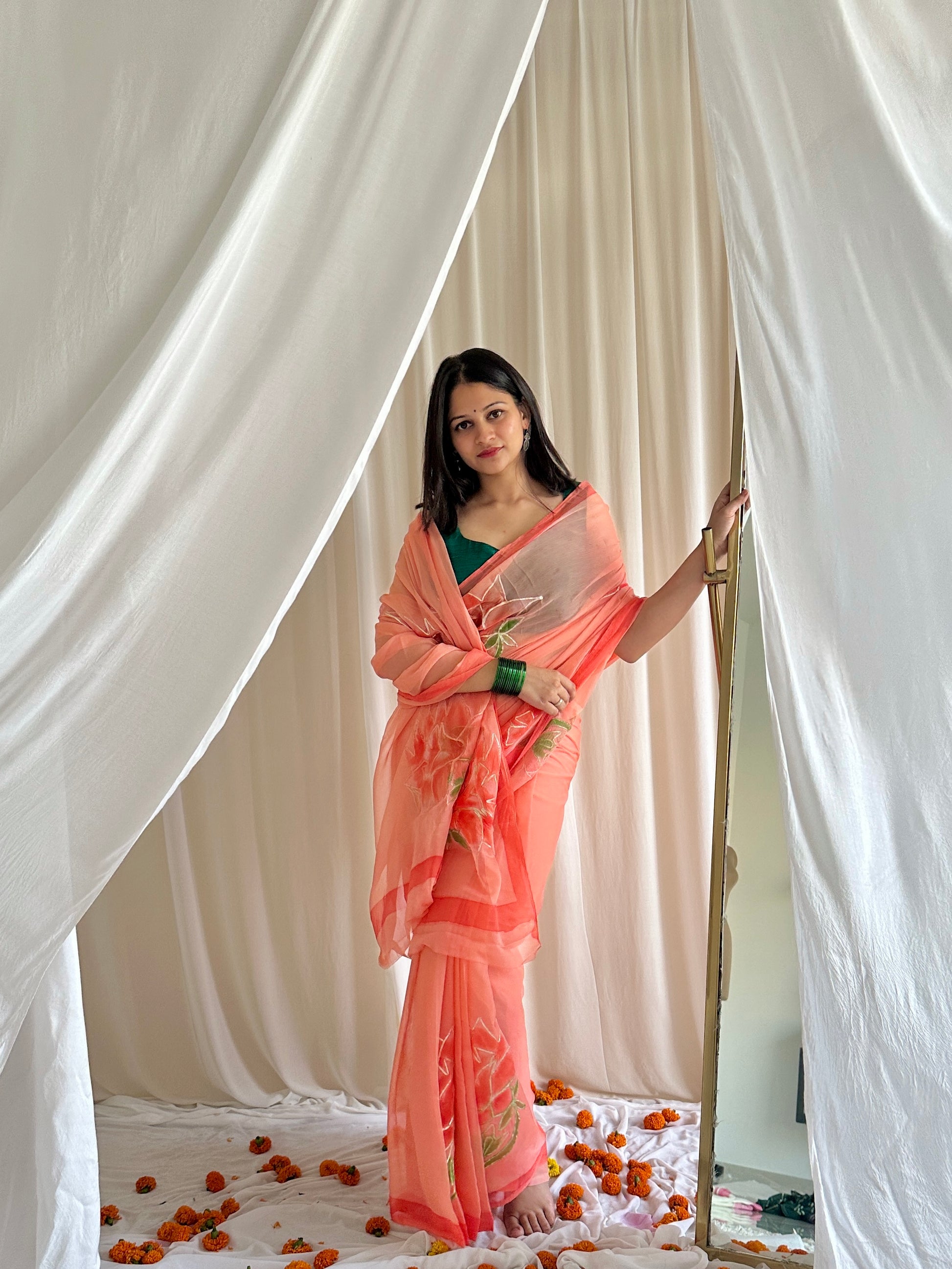 Woman in a peach saree standing between white curtains with flower petals on the floor.