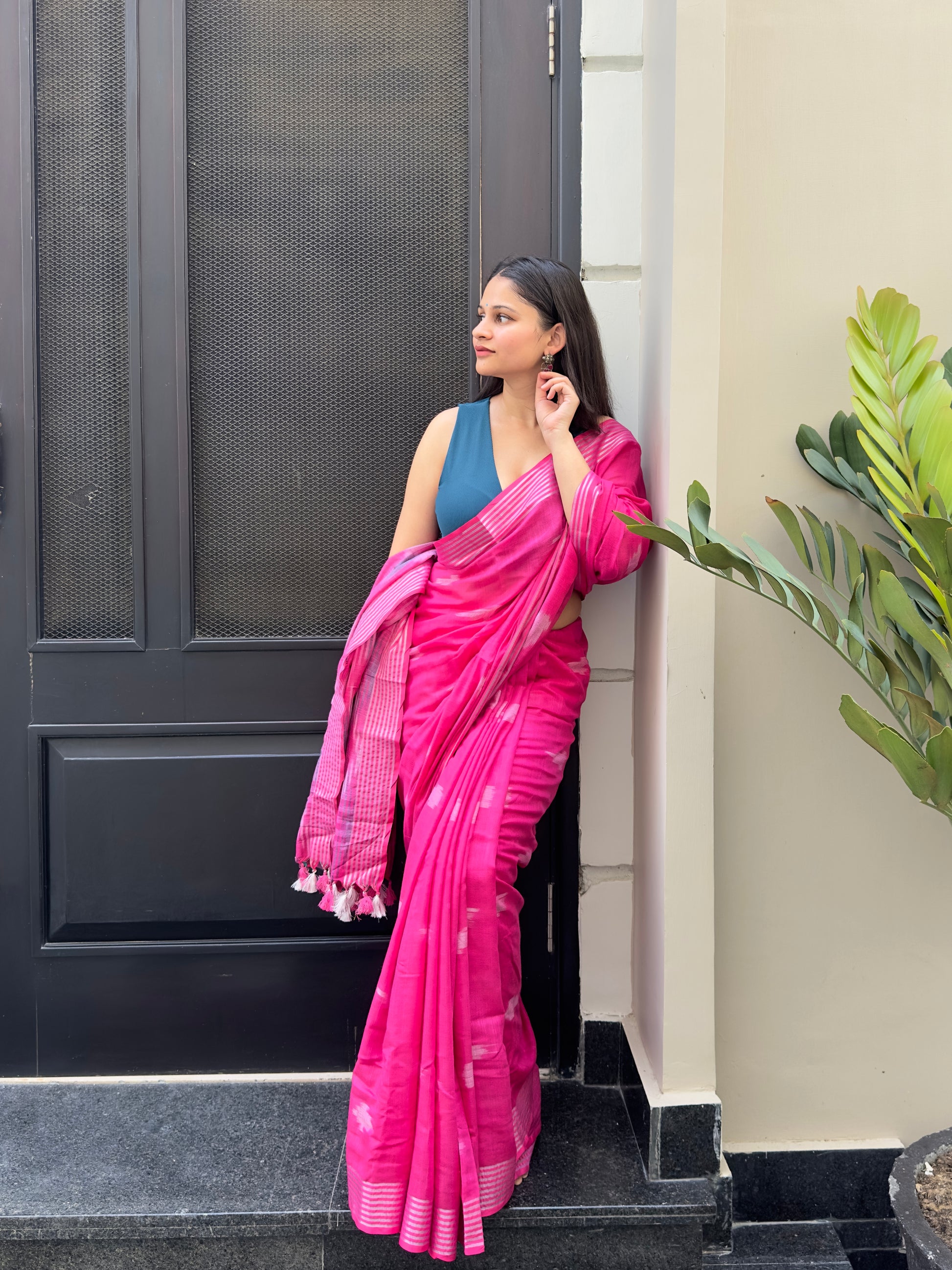 Woman in a pink saree standing against a door with a plant in the foreground