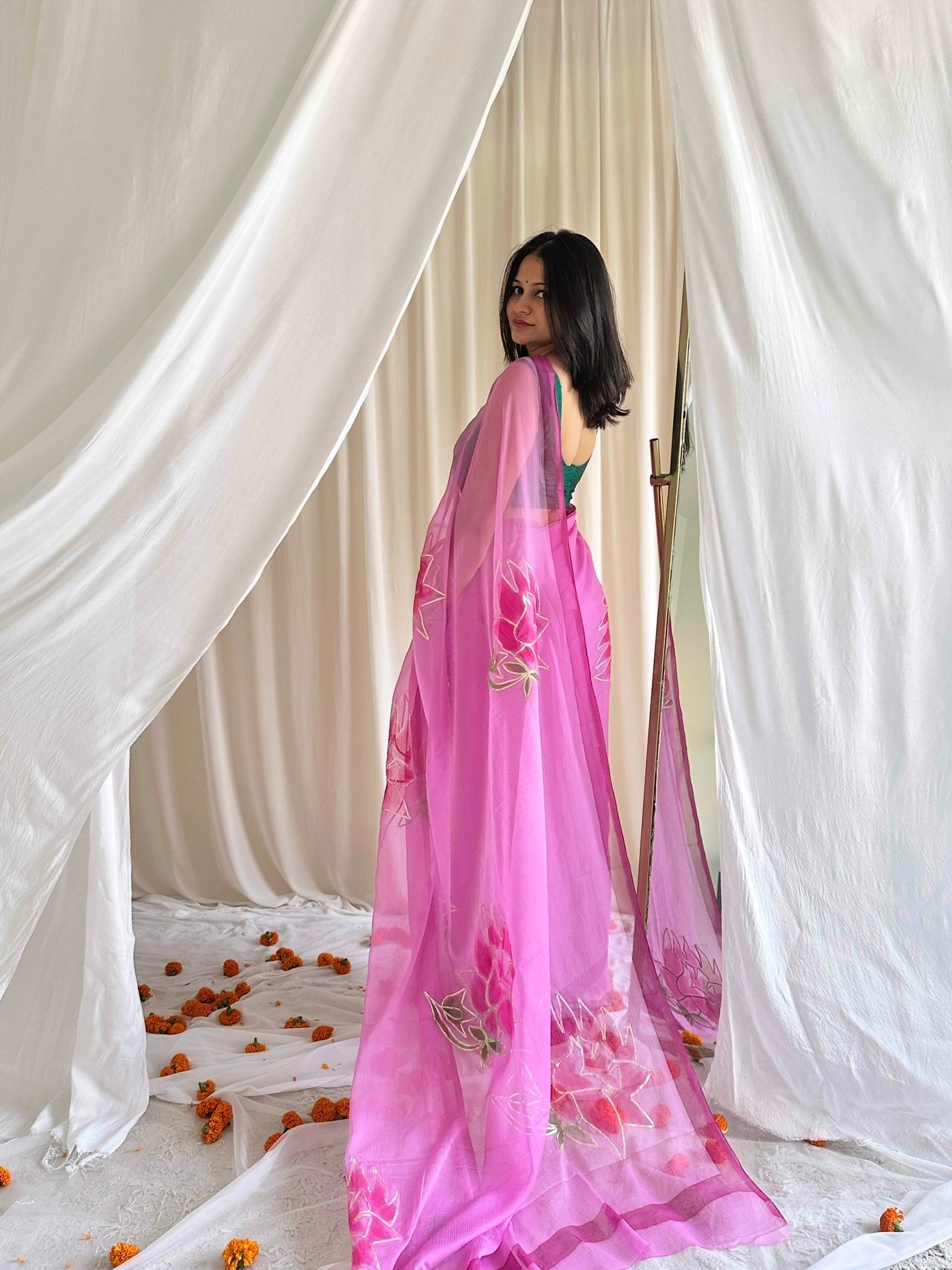 Woman wearing a pink saree with floral patterns in a draped setting.