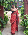 Woman in a red and black patterned saree standing in a garden.